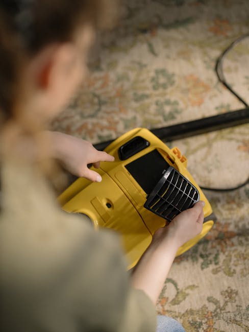 A person wearing casual clothing is kneeling on a patterned carpeted floor, using a yellow and black vacuum cleaner with a removable dust container. Their hands are inserting or removing a white cloth